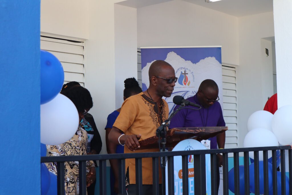 A speaker addressing an audience at a podium during an event, with blue and white balloons in the foreground and people gathered in the background.