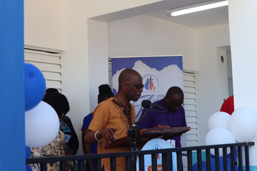 A speaker at a podium addresses an audience during an event, with balloons and a banner in the background.