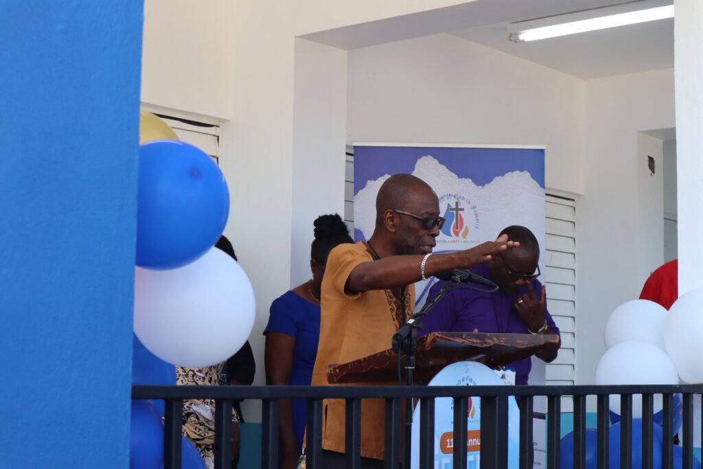 A speaker at a podium gestures while addressing an audience, with decorations in blue, white, and gold in the background.