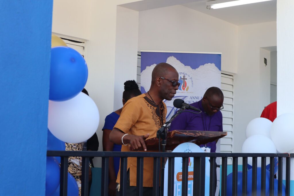 A speaker in a brown traditional shirt addresses an audience at a podium, surrounded by blue and white balloons and a backdrop with event branding.