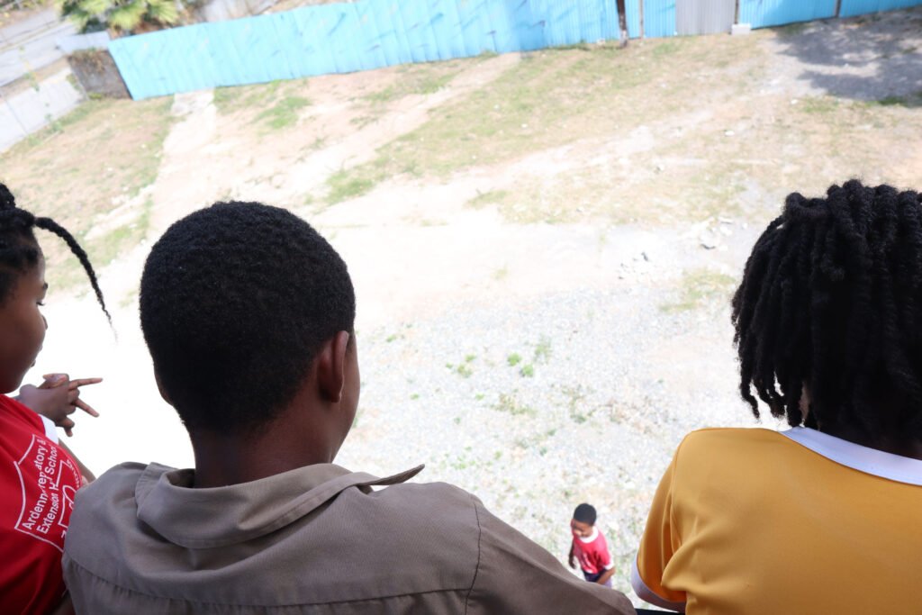 Three children watching from a height, two with short hair and one with curly hair in a yellow shirt, standing on a grassy area with a blue fence in the background.