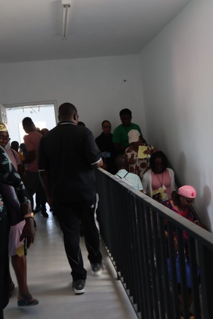 A crowded hallway with people waiting in line, some sitting against the wall and others standing. The setting appears to be indoors in a well-lit environment.