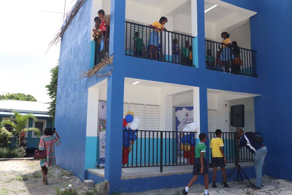 A colorful school building with multiple students gathered on the balconies, engaged in activities, and a few adults present. A person walks towards the building in the foreground.