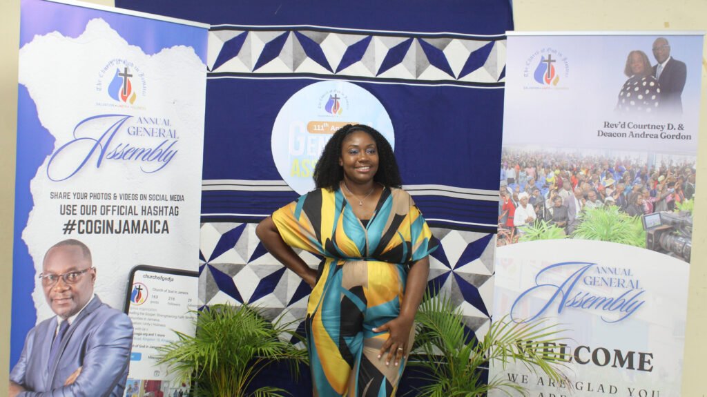 A woman in a colorful dress stands in front of banners promoting the Annual General Assembly of a church, with images of church leaders and a crowd in the background.