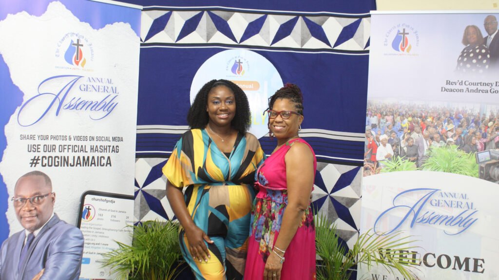 Two women posing for a photo at an event, with a backdrop displaying the words 'Annual General Assembly' and information about sharing photos on social media. One woman is wearing a colorful outfit, while the other is in a pink dress.