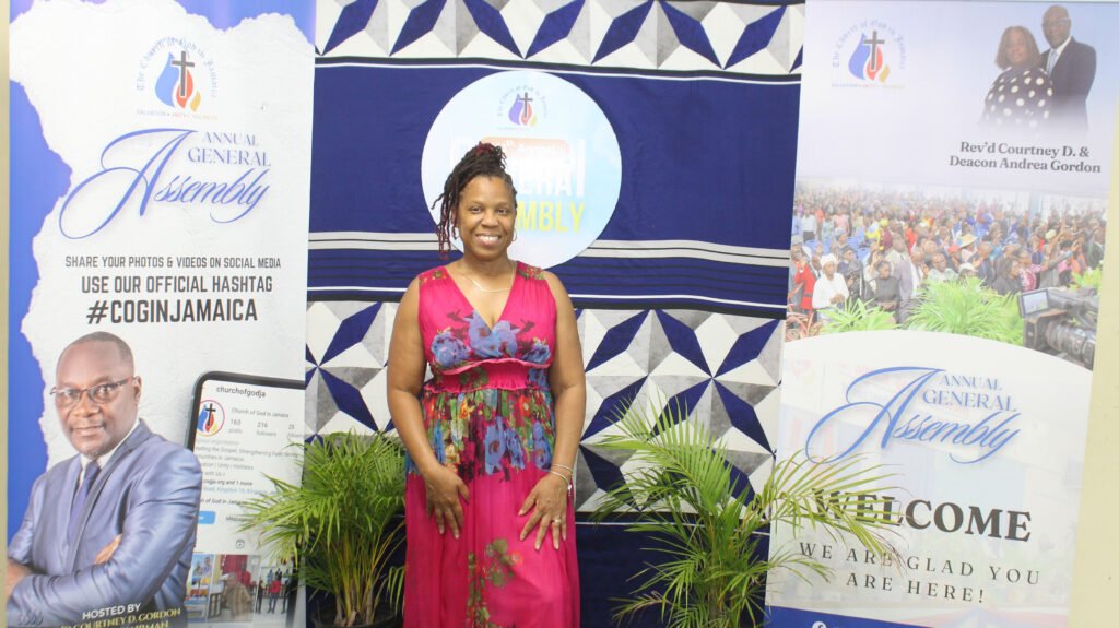 A woman in a colorful dress stands in front of a banner for the 'Annual General Assembly' event, with promotional materials and a social media hashtag.