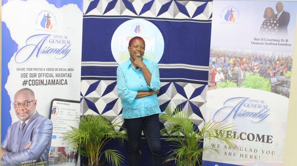 A woman smiling while standing in front of banners for the Annual General Assembly, featuring event details and a tropical plant in the foreground.