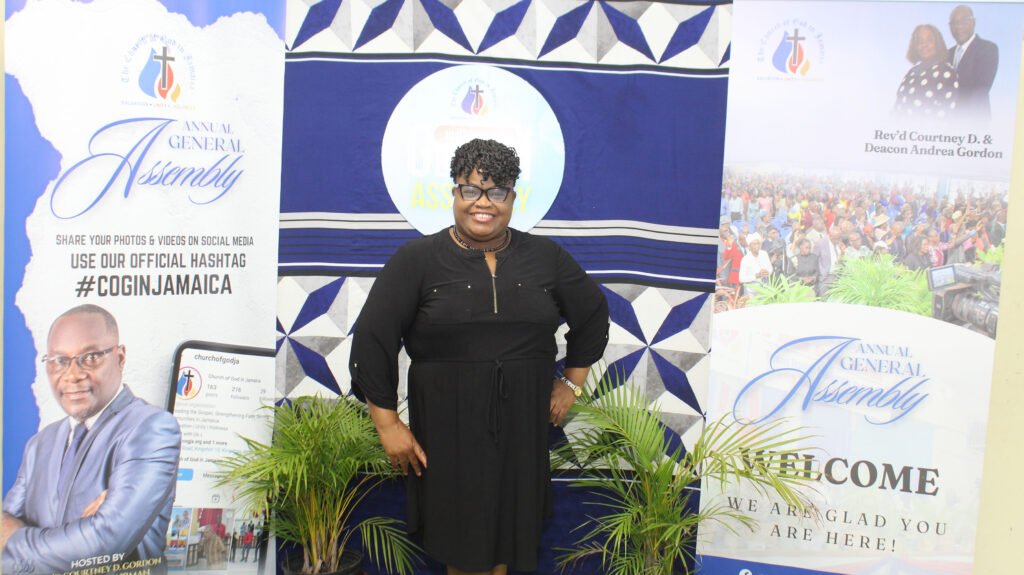 A woman standing in front of a banner for the Annual General Assembly, surrounded by decorative elements and plants.