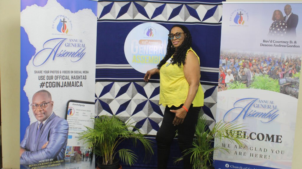A woman in a bright yellow top stands next to banners for the Annual General Assembly, featuring event branding and a photo of a large crowd. The setting is decorated with green plants and the event hashtag #COGINJAMAICA.