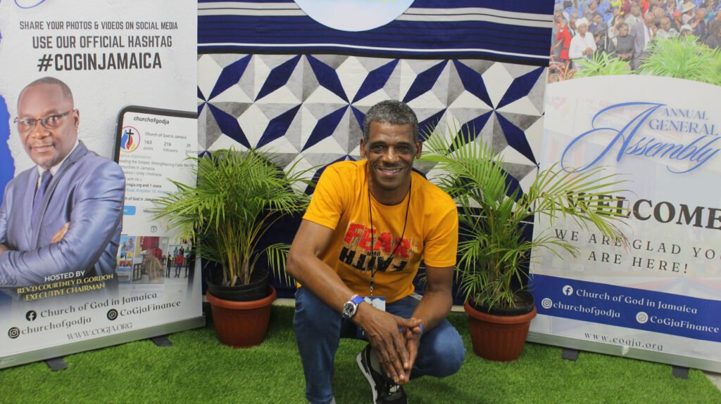 A man in an orange shirt poses in front of two banners at an event, with plants in the foreground and a geometric patterned backdrop.