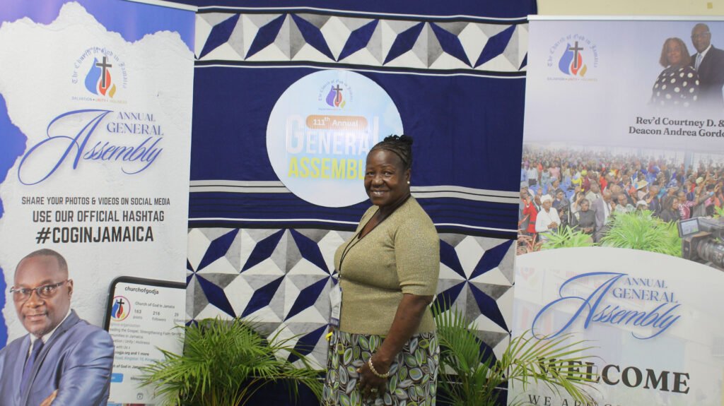 A woman smiling at a photo backdrop displaying the logo and details of the Annual General Assembly event. The backdrop features colorful designs and messages about sharing photos and videos on social media using the hashtag #COGINJAMAICA.