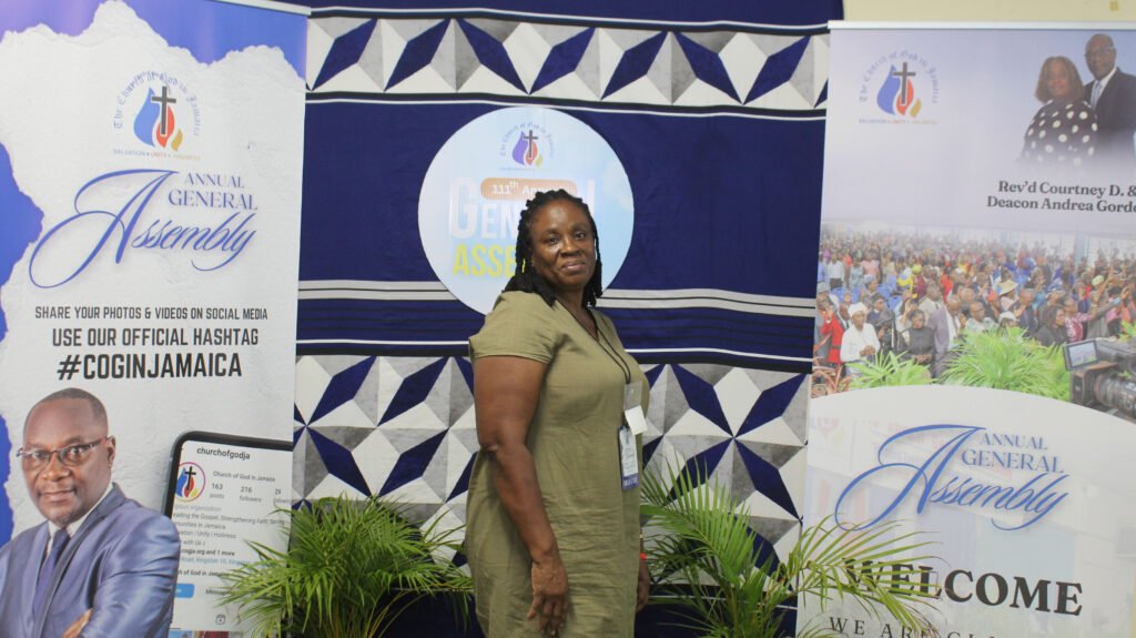 A woman stands in front of banners promoting an annual general assembly, featuring a graphic design and notifying attendees to share their photos and videos on social media using a specific hashtag.