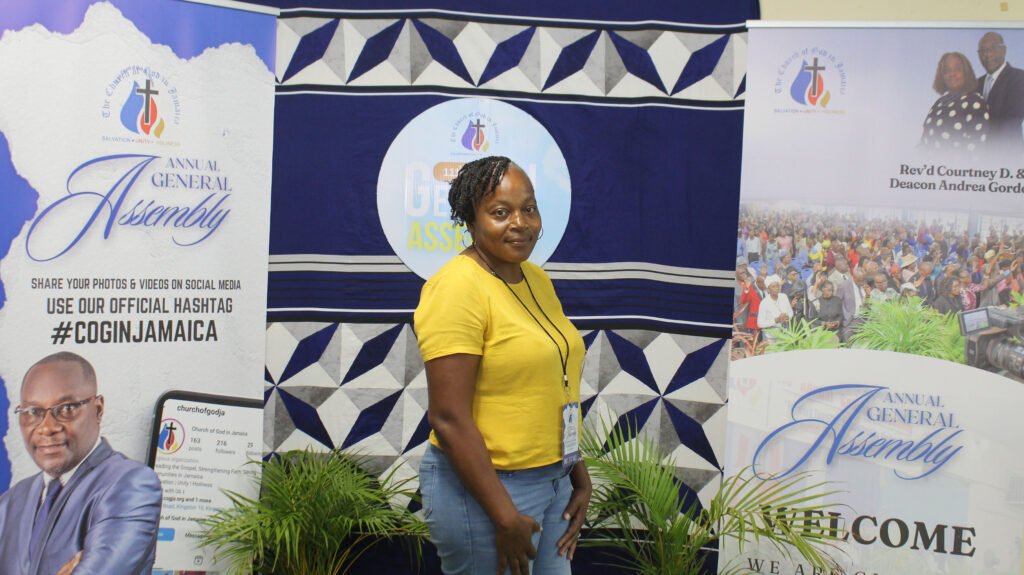 A woman in a yellow shirt stands in front of a backdrop featuring banners for an Annual General Assembly event, with images of church leaders and an audience.