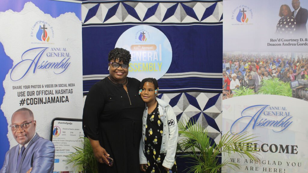 A woman and a child posing together in front of a backdrop featuring banners for an annual general assembly event.