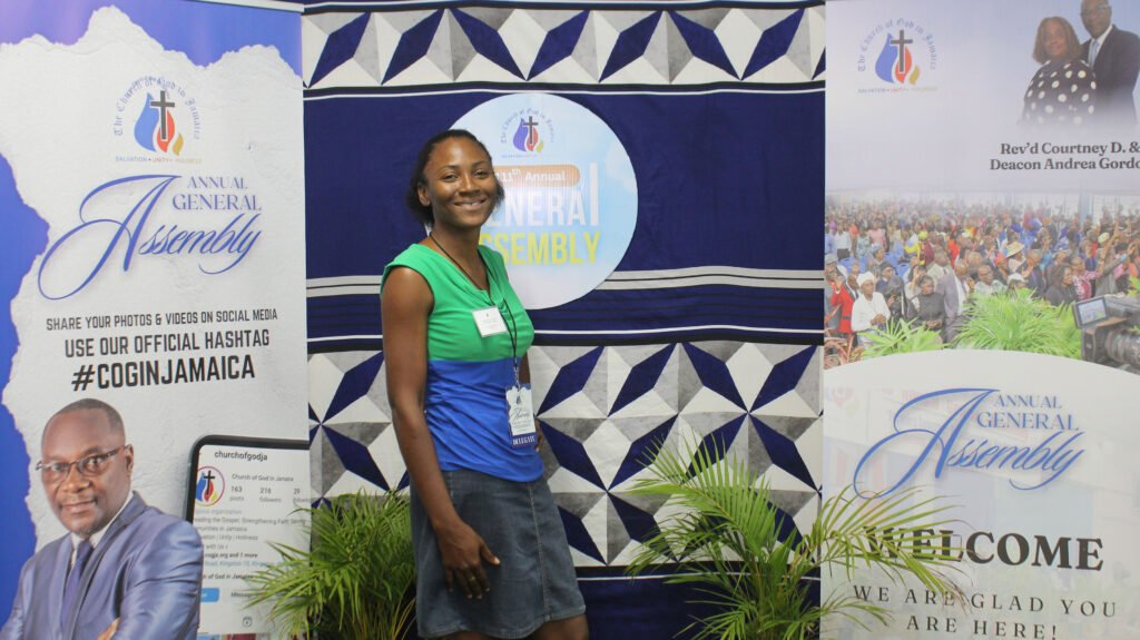 A person smiling at an event backdrop featuring the words 'Annual General Assembly' and promotional graphics. The backdrop includes images of speakers and a congregation.