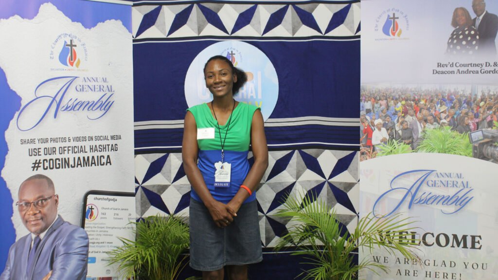 A woman stands in front of a backdrop at an event, wearing a green top and denim skirt, smiling for the camera. The backdrop features text promoting the Annual General Assembly and social media engagement.