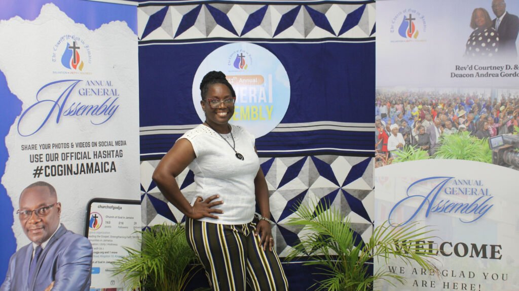 A woman standing in front of a backdrop featuring promotional materials for an annual general assembly, smiling and posing with her hand on her hip.