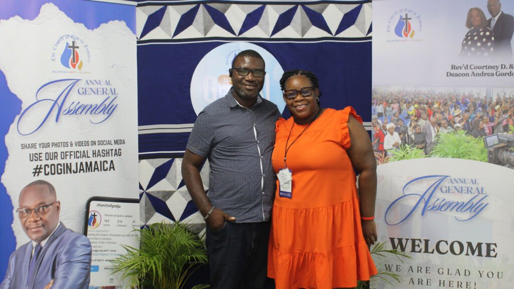 A man and a woman pose together at an event, smiling, with banners promoting the Annual General Assembly in the background.