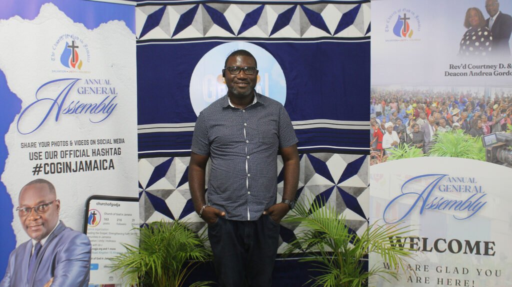 A man poses in front of a backdrop featuring the text 'Annual General Assembly' and promotional materials for a church event, with imagery of attendees in the background.