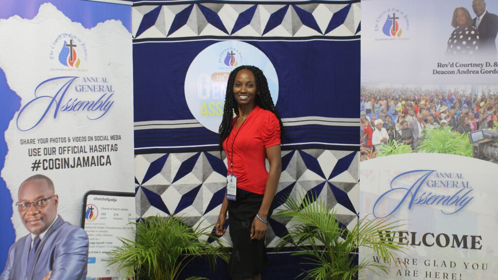 A woman in a red shirt stands at an event backdrop for an Annual General Assembly, featuring promotional graphics and a gathering of people in the background.