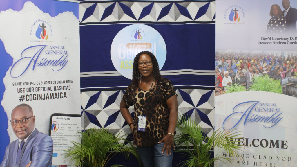 A woman standing in front of a display for the Annual General Assembly of the Church of God in Jamaica, featuring promotional materials and a logo, with attendees visible in the background.