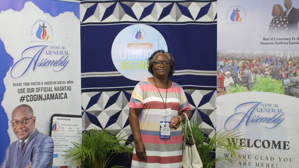 A woman with glasses stands in front of banners at a church event, smiling. The banners display information about the Annual General Assembly and include images of church leaders and a crowd.