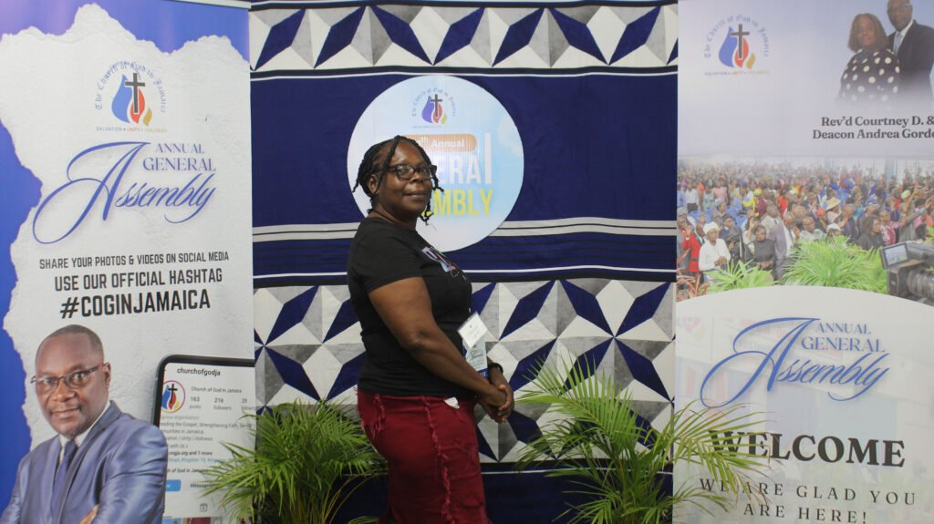 A woman standing in front of a backdrop displaying banners for the Annual General Assembly of the Church of God in Jamaica, with graphics and text emphasizing the event and social media engagement.