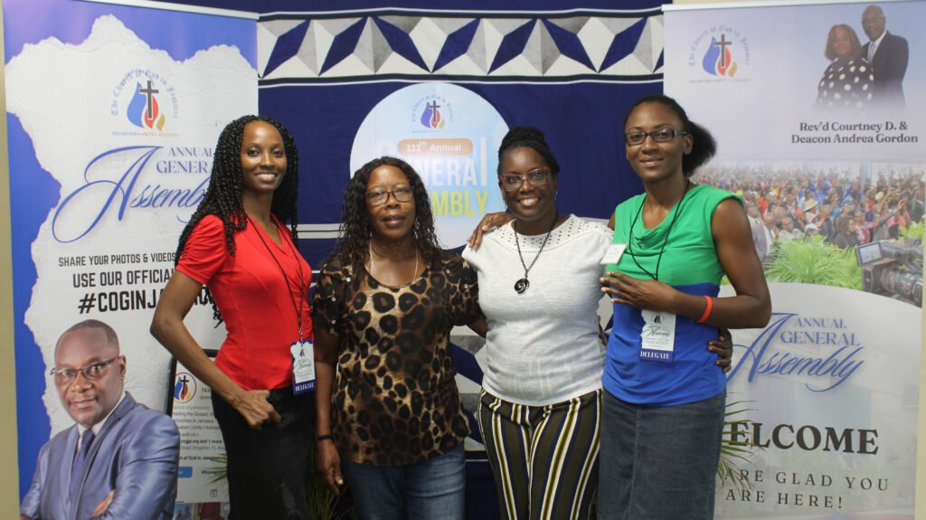 A group of four women posing together at an event, standing in front of a banner that reads 'Annual General Assembly.' The women are smiling, dressed in a variety of outfits, and the background features additional event branding.