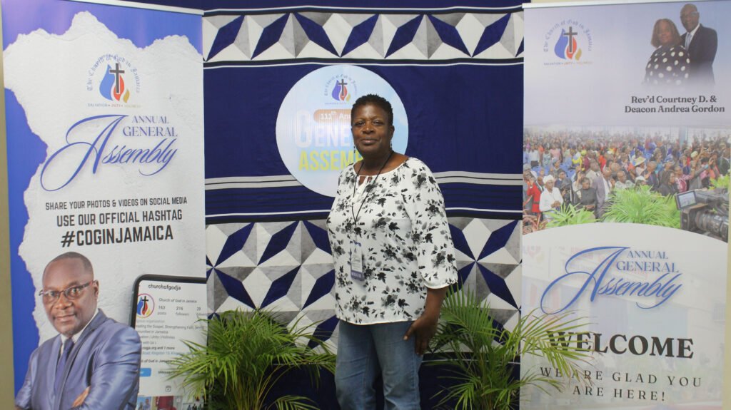 A woman stands in front of banners for an Annual General Assembly event, surrounded by green plants.