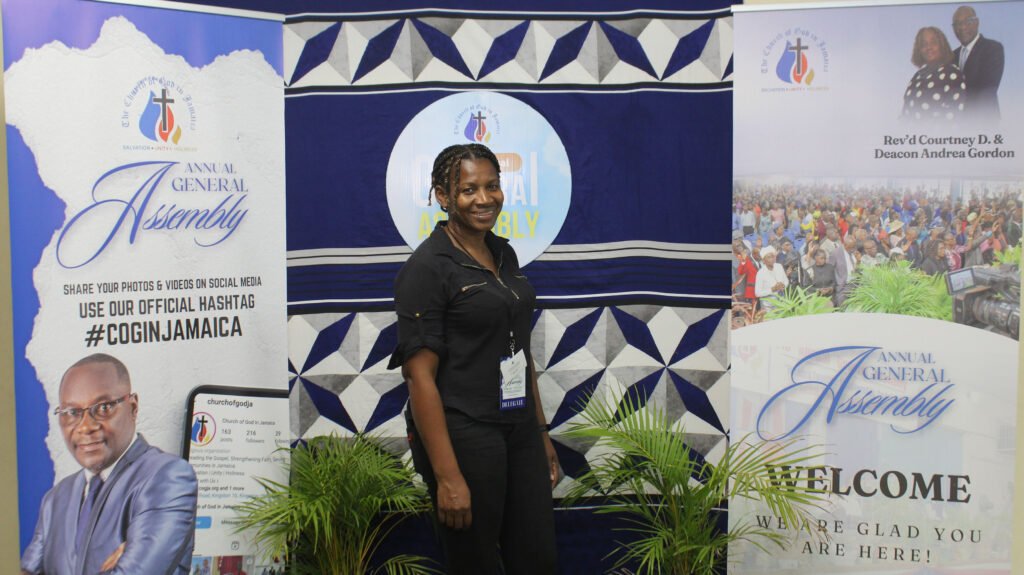 A woman stands smiling in front of banners for an Annual General Assembly event, with details about sharing photos and videos on social media.