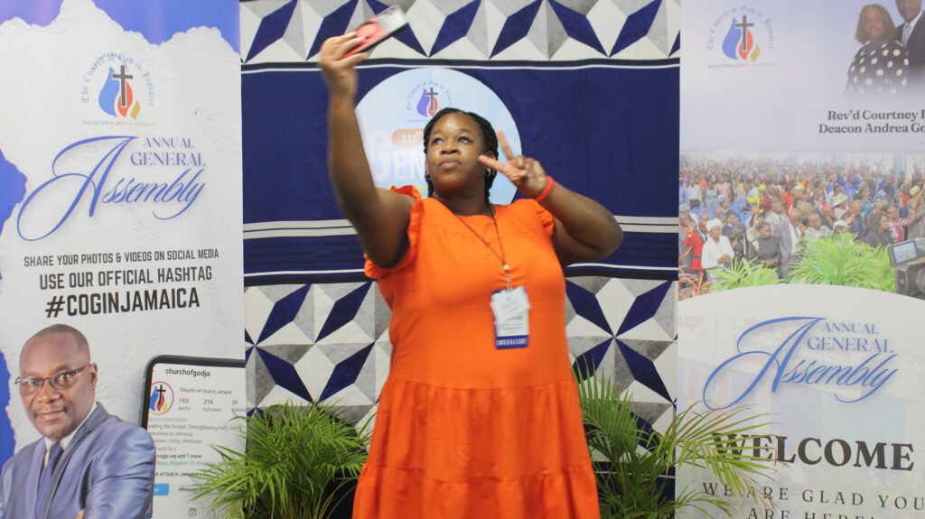 A woman in an orange dress takes a selfie at a church event, surrounded by promotional banners for the Annual General Assembly and social media engagement.