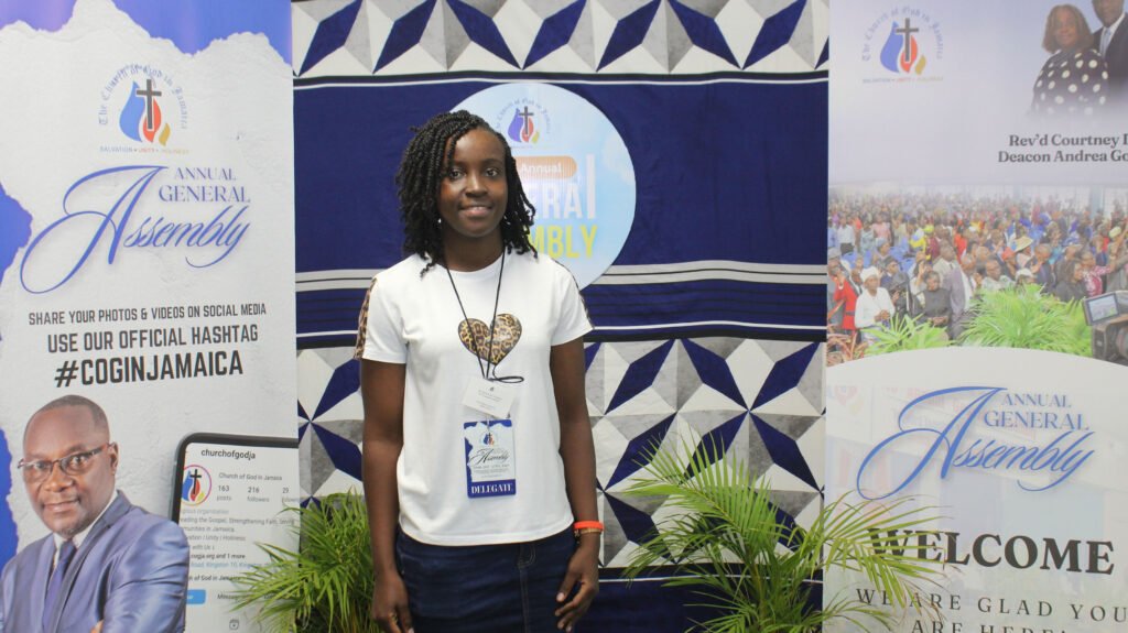 A young woman stands in front of banners promoting the Annual General Assembly event, wearing a white shirt with a heart design. The background features blue and white decor, illustrating the ambiance of the gathering.