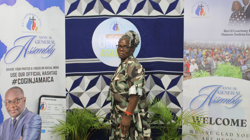 An elderly woman in a camouflage outfit stands in front of banners for the Annual General Assembly of the Church of God in Jamaica, featuring a blue and white backdrop with the event's logo.