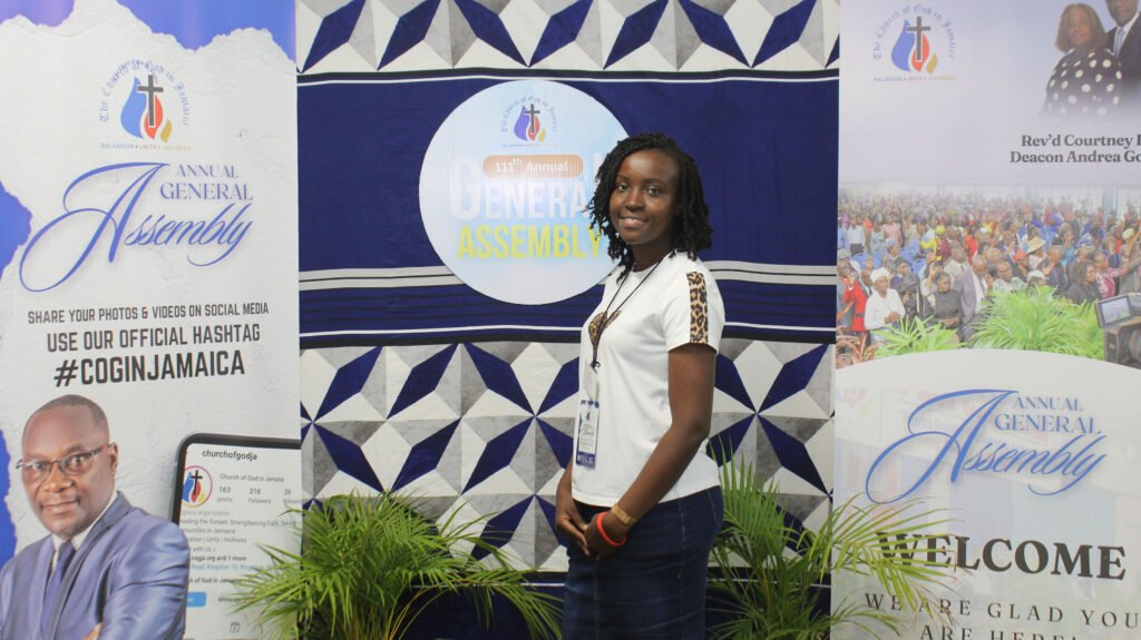 A woman stands smiling next to banners promoting the Annual General Assembly of a church, featuring the church logo and information about sharing photos on social media.