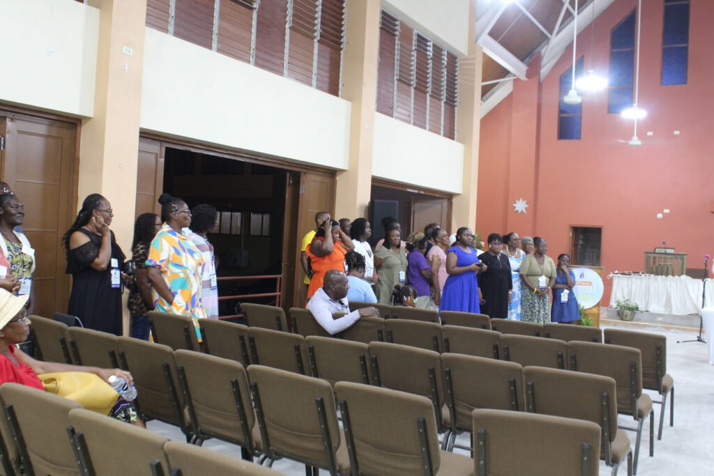 A group of women standing in a church setting, some wearing colorful dresses, while others are in more formal attire. The background features wooden doors and a bright orange wall.