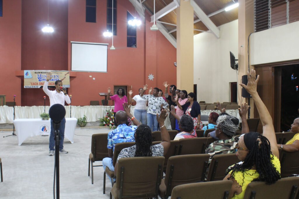 A speaker stands at the front of a church, gesturing enthusiastically as the audience raises their hands in response. Several attendees are seated, while others stand nearby, showing engagement in the gathering.