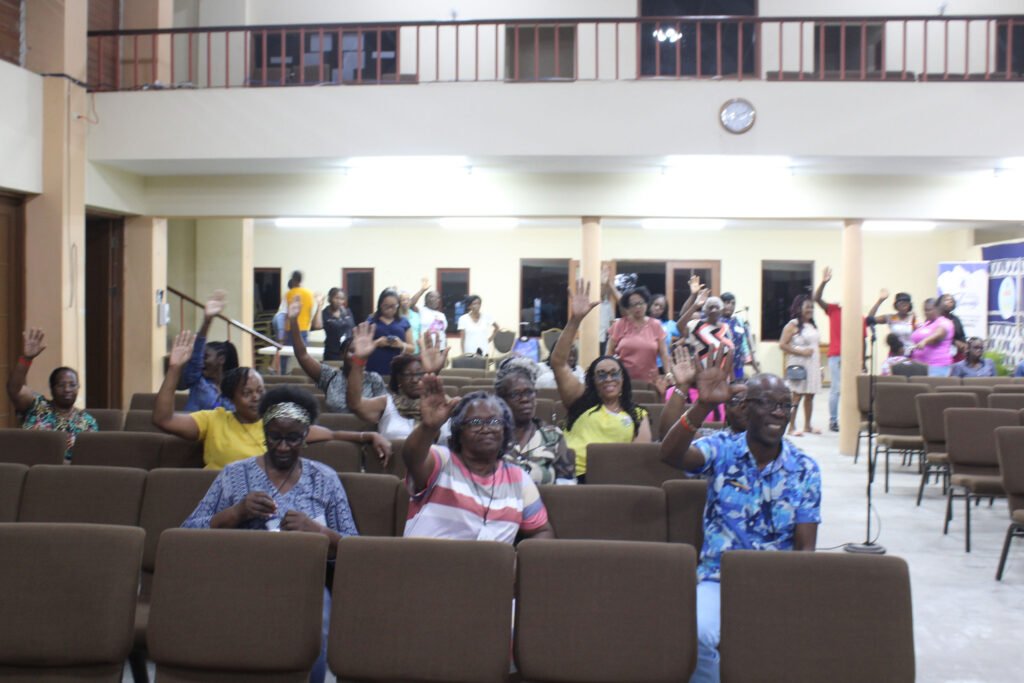 A group of people in a hall raising their hands, sitting in chairs. Some individuals are smiling and engaging with the audience. The background shows more attendees and an indoor setting.
