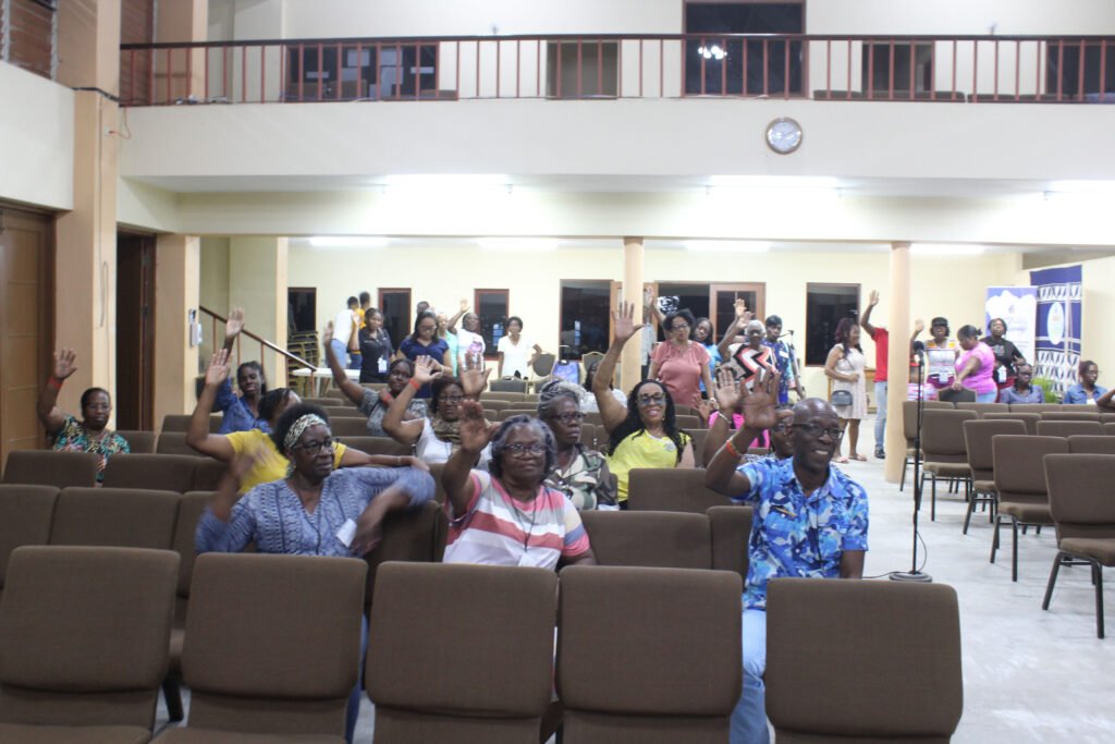 A group of people seated in a hall, many of them raising their hands, indicating participation or enthusiasm, with empty chairs in front and a few individuals milling about in the background.