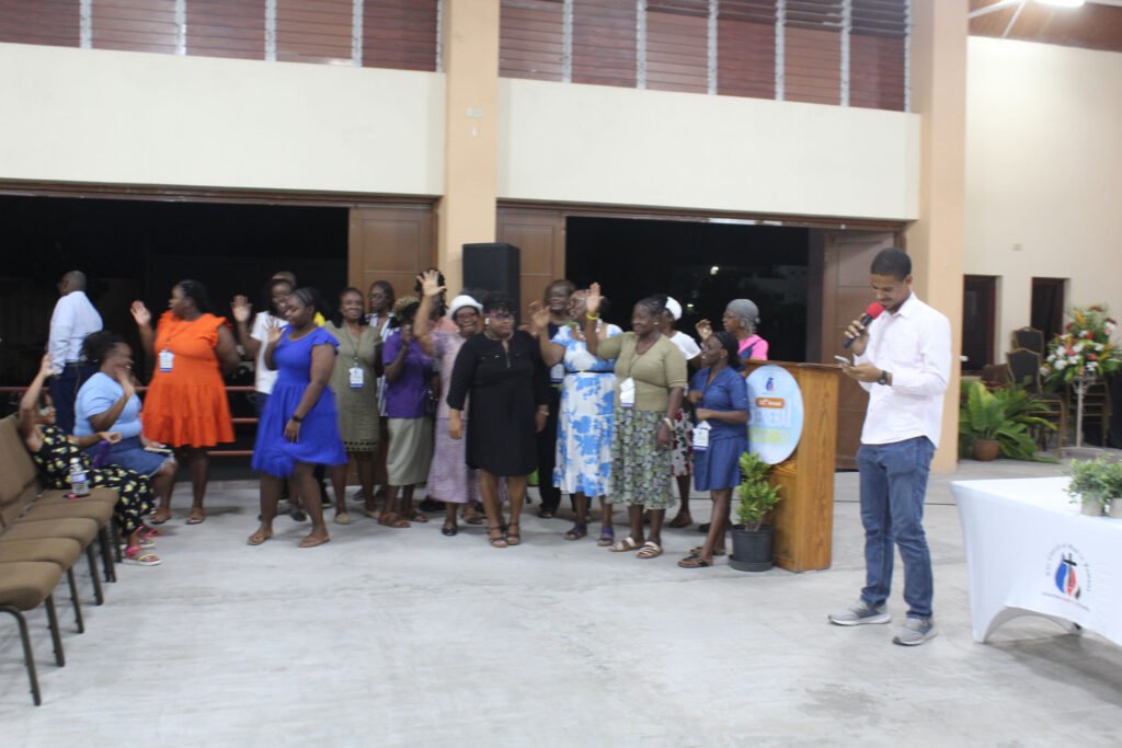 A group of people, mostly women, are gathered in a spacious venue, with some waving and others standing together while one person at a podium speaks. Decorations and seating are visible in the background.