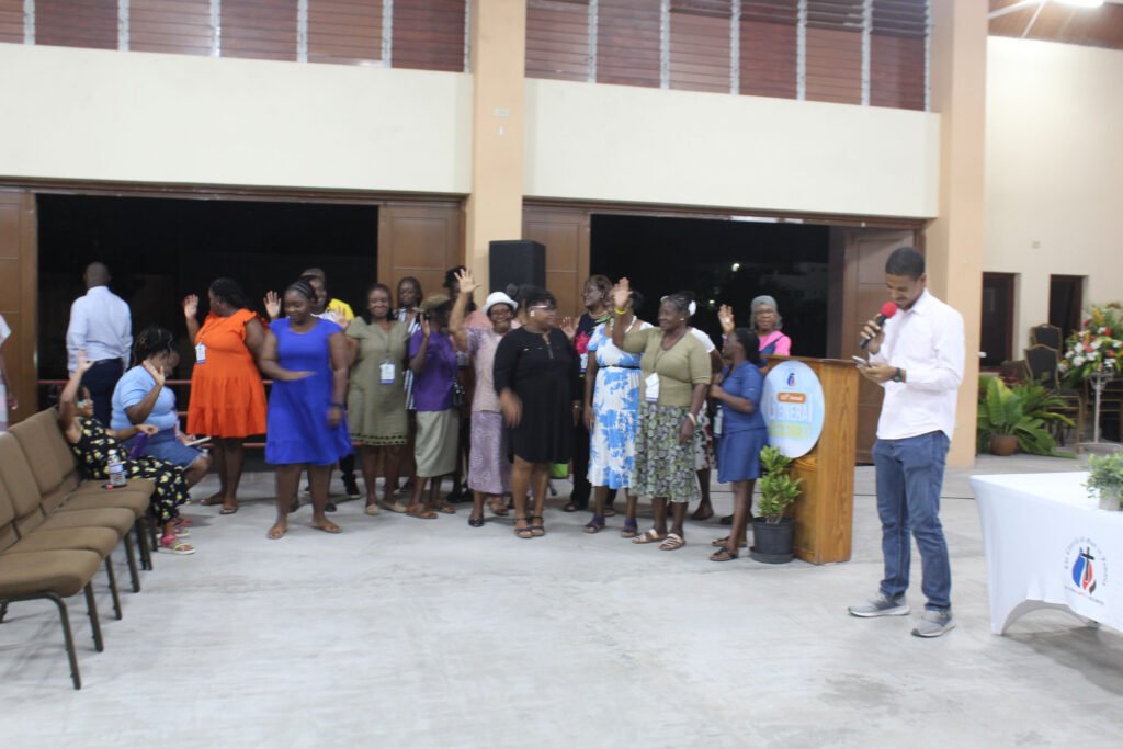 A group of women standing and celebrating during a gathering, with one person speaking at a podium. Some participants are raising their hands, while others sit on chairs in the foreground.