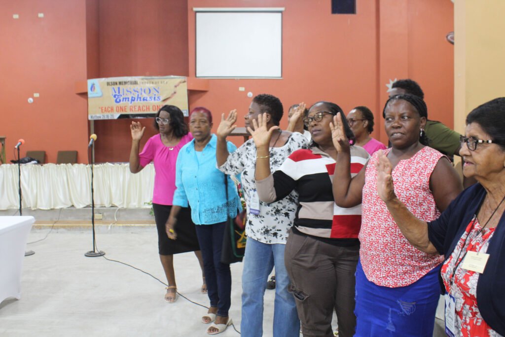 A group of women standing together with raised hands during a ceremony, with a banner in the background that reads 'Mission Emphasis' in a church setting.