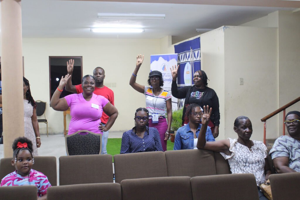 A group of people raising their hands in a community meeting setting, with a mix of adults and a child. The atmosphere is lively and engaged.