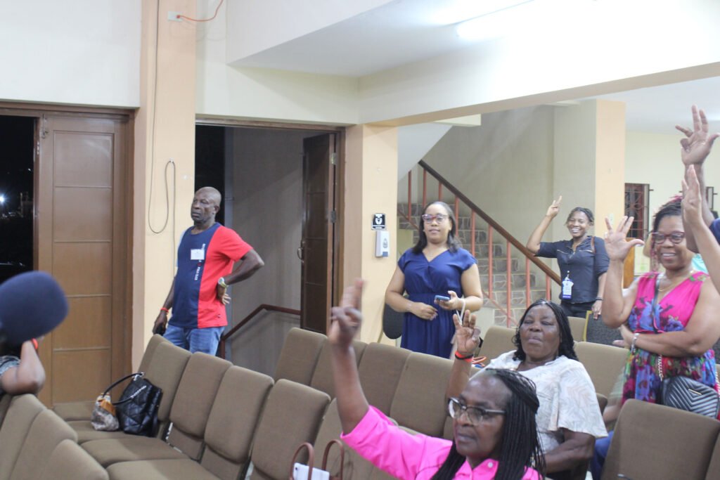 A group of people in a meeting, some raising their hands, expressing engagement. The setting has brown chairs and a light-colored wall, with a staircase visible in the background.