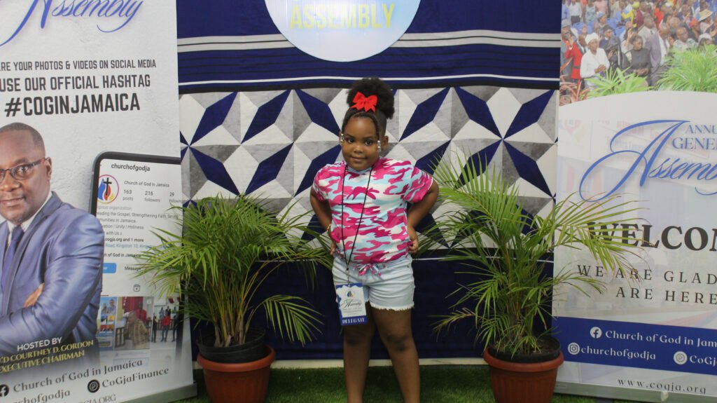A young girl wearing a pink camouflage shirt and shorts standing in front of a decorative backdrop with plants on either side. The backdrop features banners and information related to a church event.