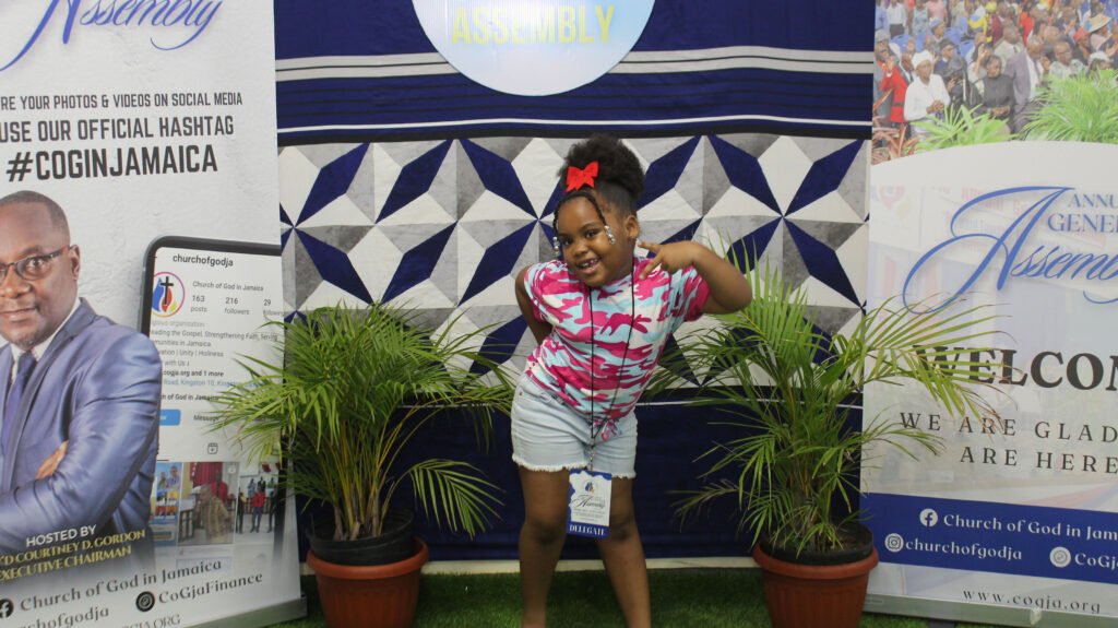 A young girl with a playful pose, wearing a pink shirt and denim shorts, stands in front of a decorated backdrop featuring tropical plants and banners from a church event.