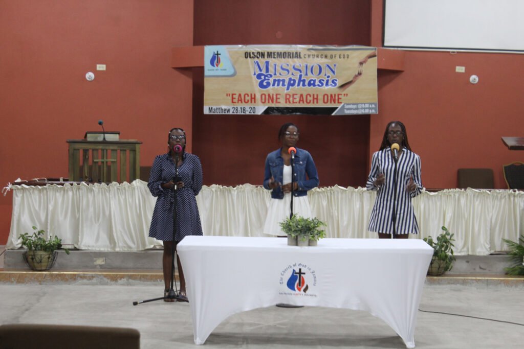 Three women singing on stage in a church setting, with a banner behind them that reads 'OLSON MEMORIAL CHURCH OF GOD' and 'MISSION EMPHASIS'.