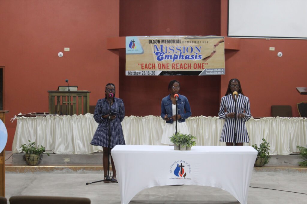 Three women singing at a church event, standing behind a table with a white tablecloth and microphones, in front of a banner that reads "Mission Emphasis".