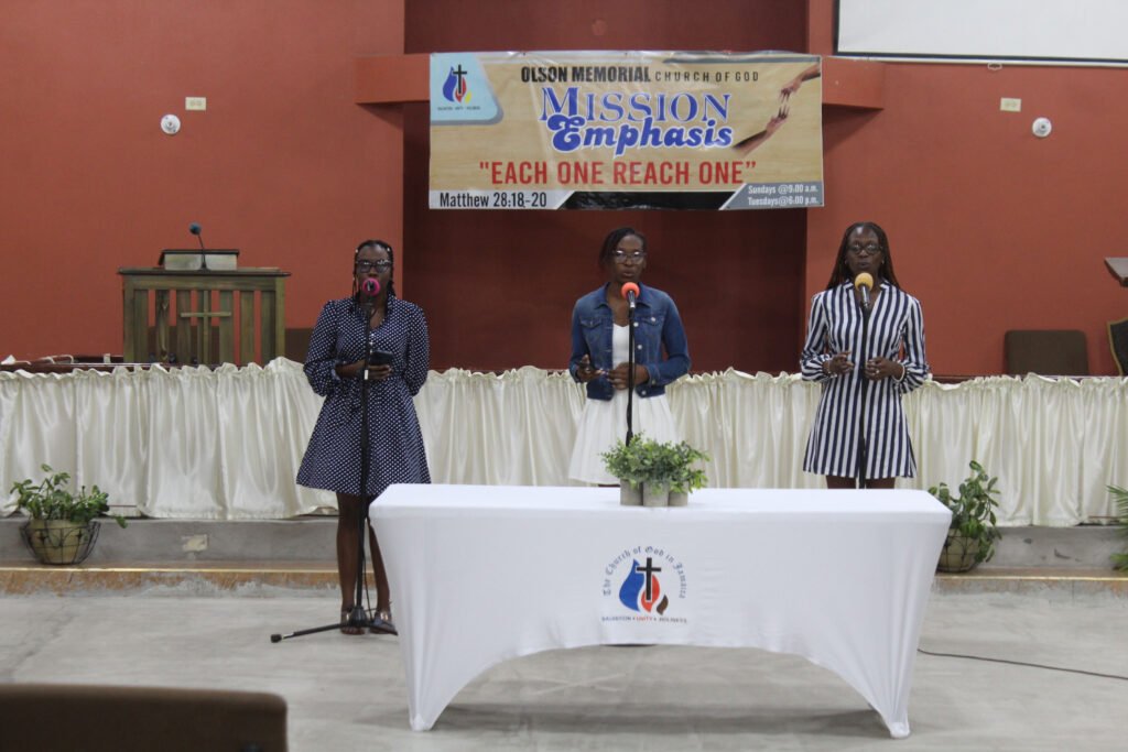 Three women singing at a church service, each with a microphone, standing in front of a podium and a banner with a biblical message.