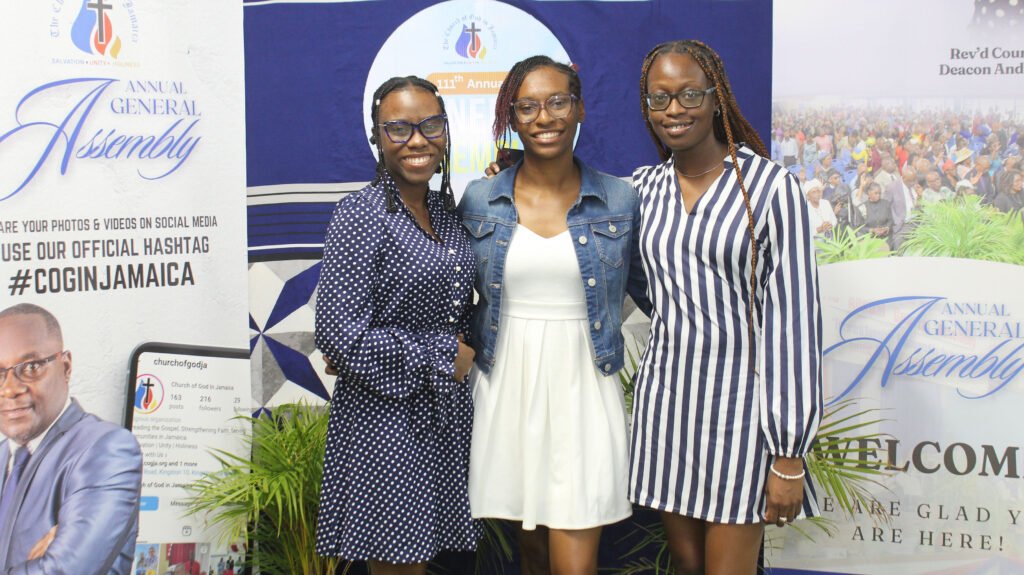Three women posing together at an event, smiling. The backdrop features the text 'Annual General Assembly' and the hashtags for social media engagement.