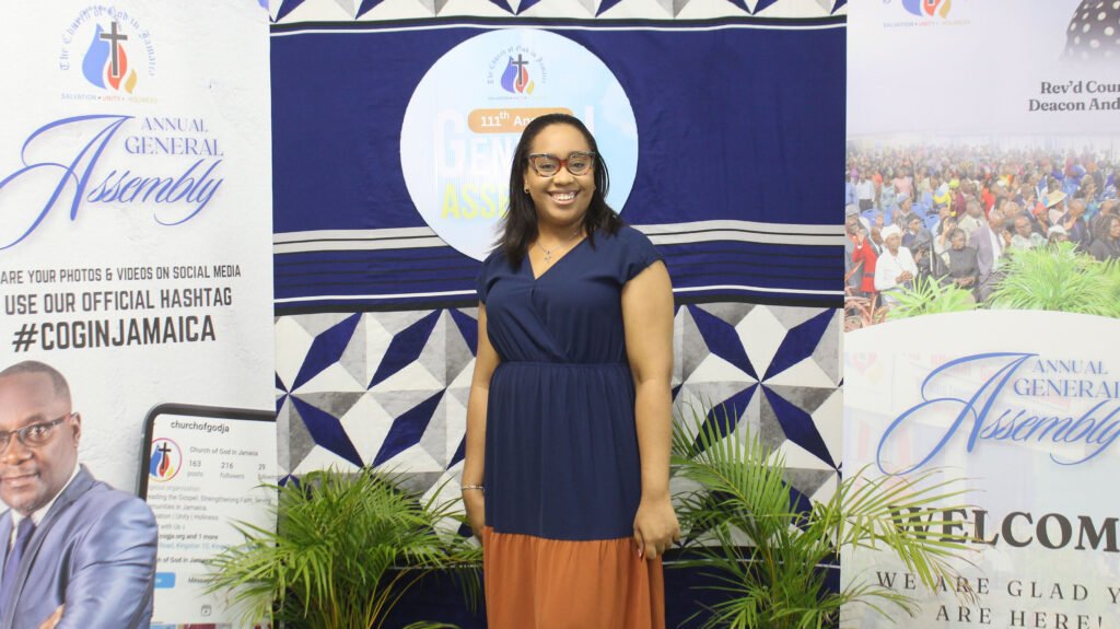 A woman standing in front of a backdrop at the Annual General Assembly, featuring the event branding and social media hashtags.